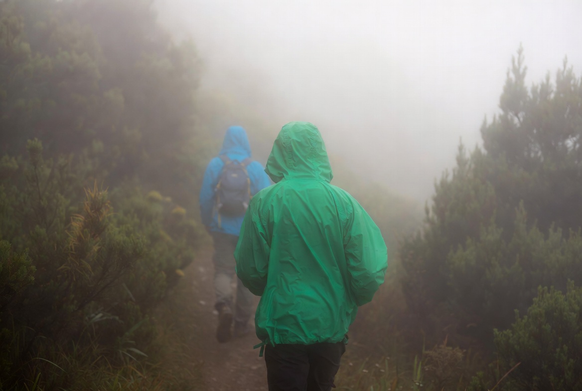 Two hikers wearing light rain shells on a misty trail, one in a green shell and one in a blue shell